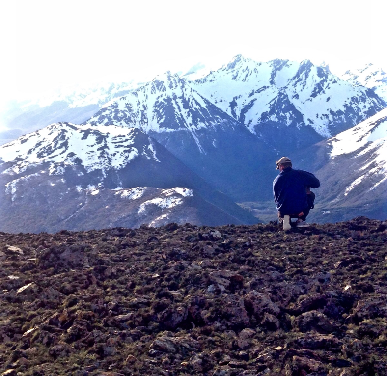 A person crouches on a rocky ridge looking out over snow-capped mountain ranges, blue and white tones, peaceful expansive view.