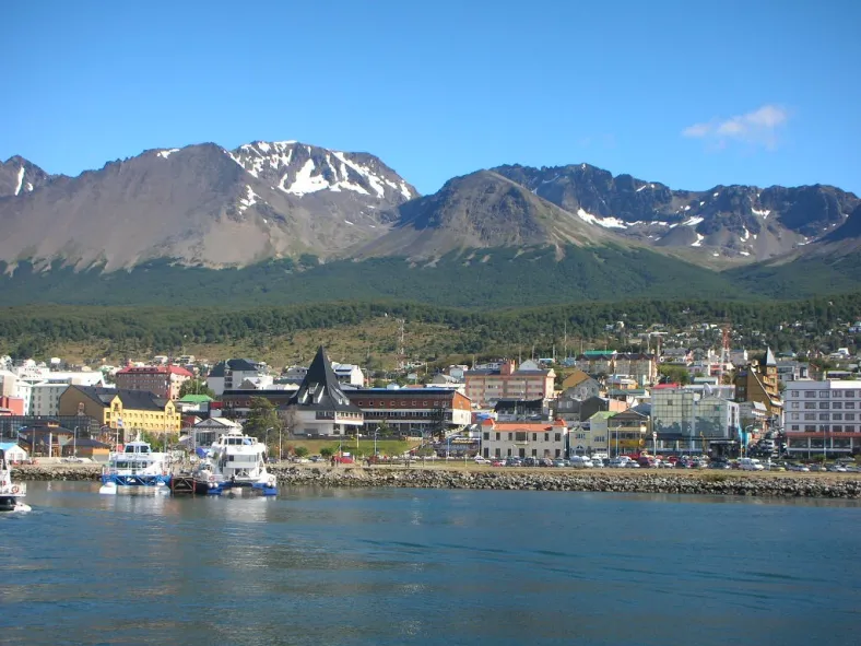 A coastal town with colorful buildings nestled at the base of snow-capped mountains, viewed from the water with boats in the foreground under soft daylight.